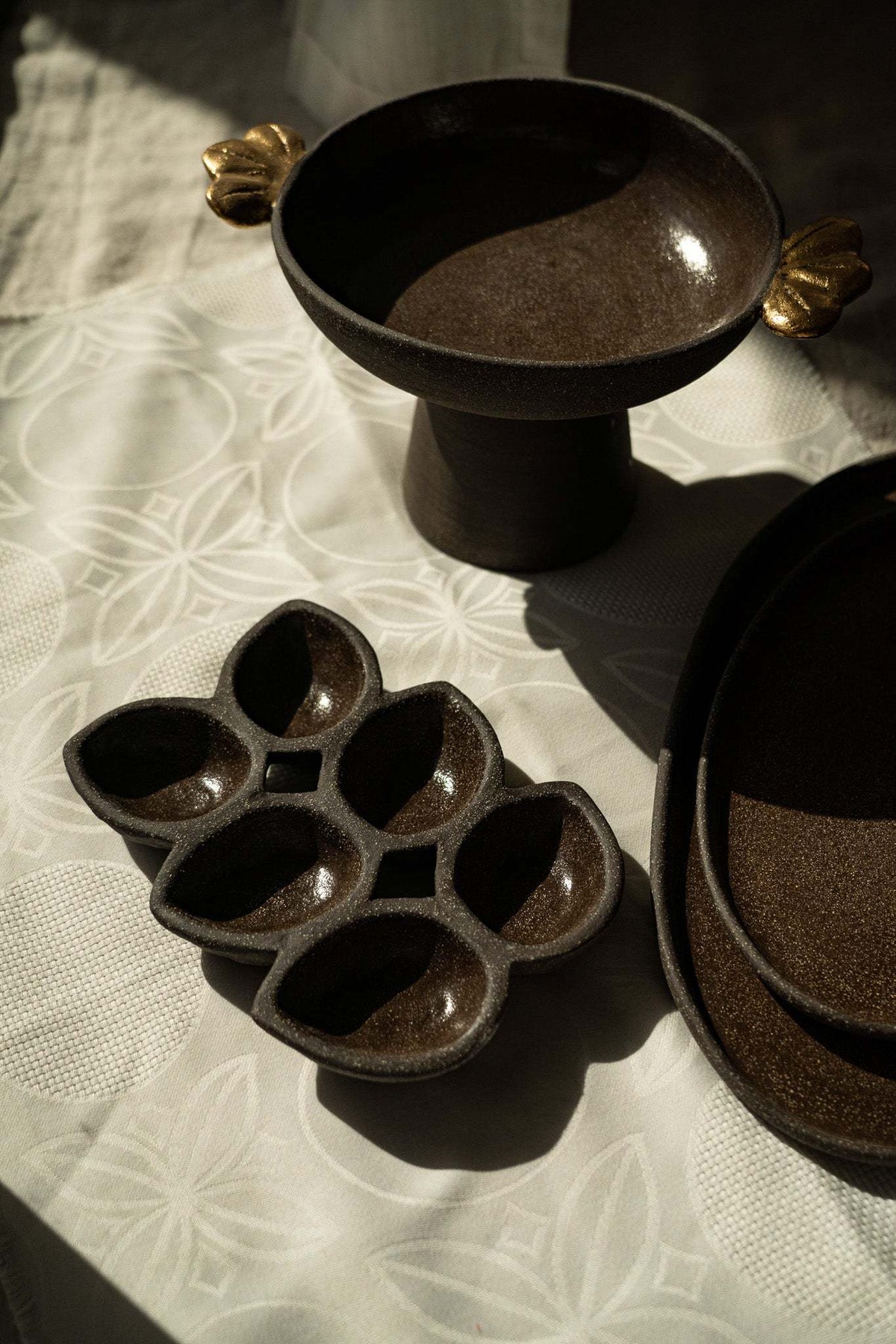 Stoneware pedestal fruit bowl in dark brown with a smooth glazed interior and gold leaf-shaped handles.