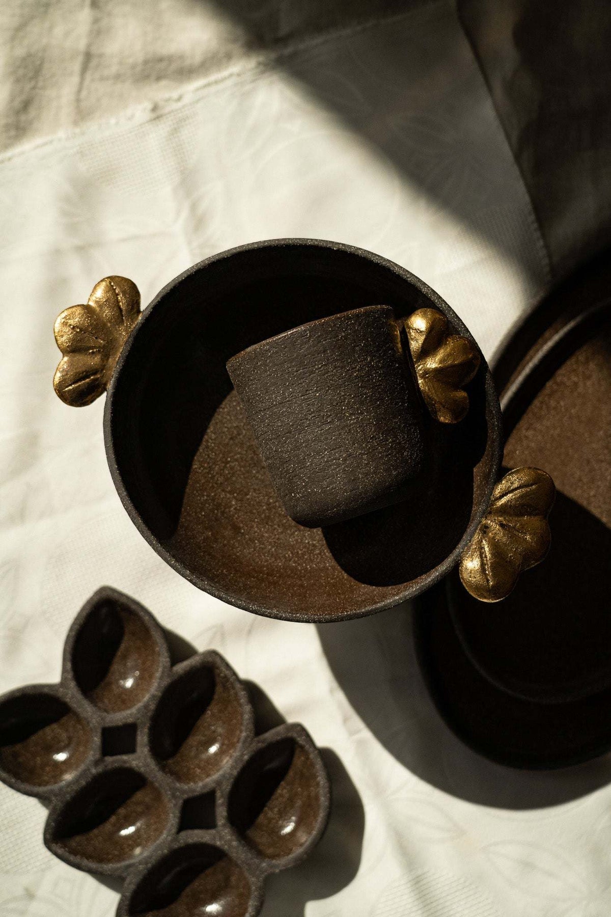Stoneware pedestal fruit bowl in deep charcoal-brown with a textured finish and sculptural gold leaf accents on the rim.