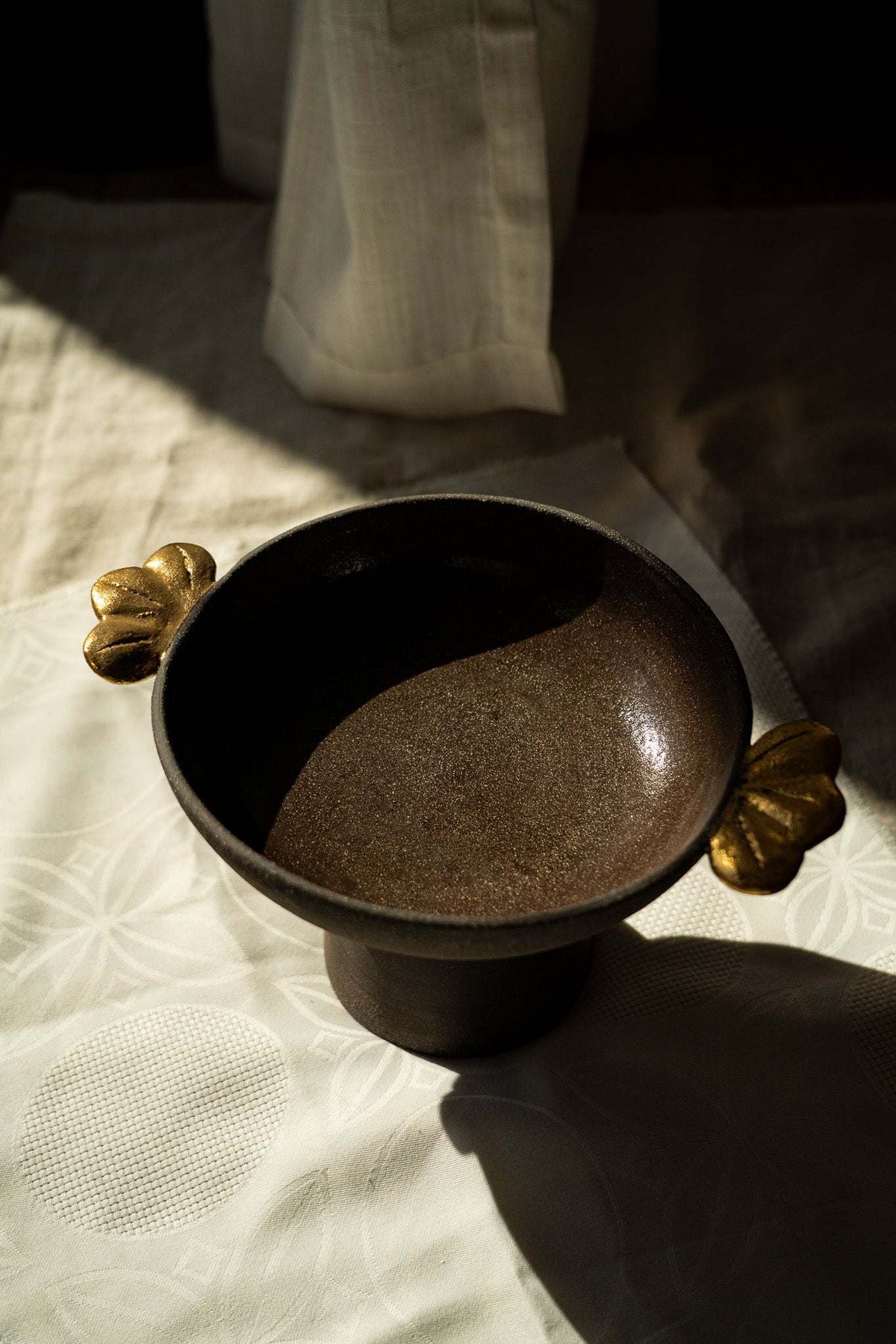 Pedestal fruit bowl in dark brown textured stoneware, with a smooth interior and sculpted gold leaf handles on both sides.