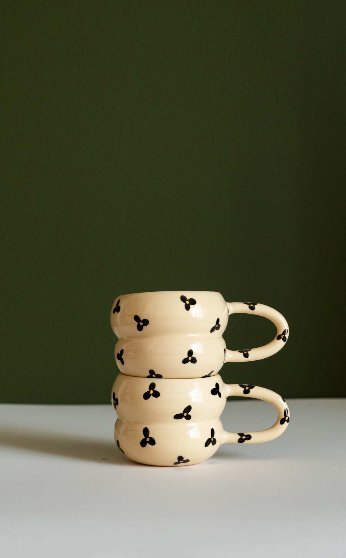 Bubble Mug in glossy cream ceramic, with a sculptural double-ring shape, curved handle, and scattered black three-petal flowers with yellow centers.