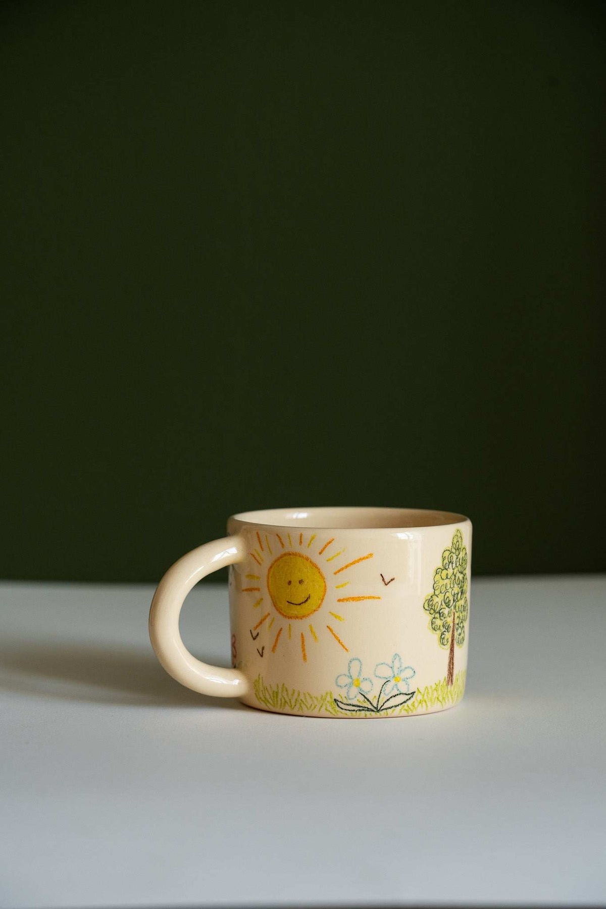 Kids Artwork Stoneware Mug in cream ceramic, decorated with a hand-drawn smiling sun, green tree, flowers and birds.