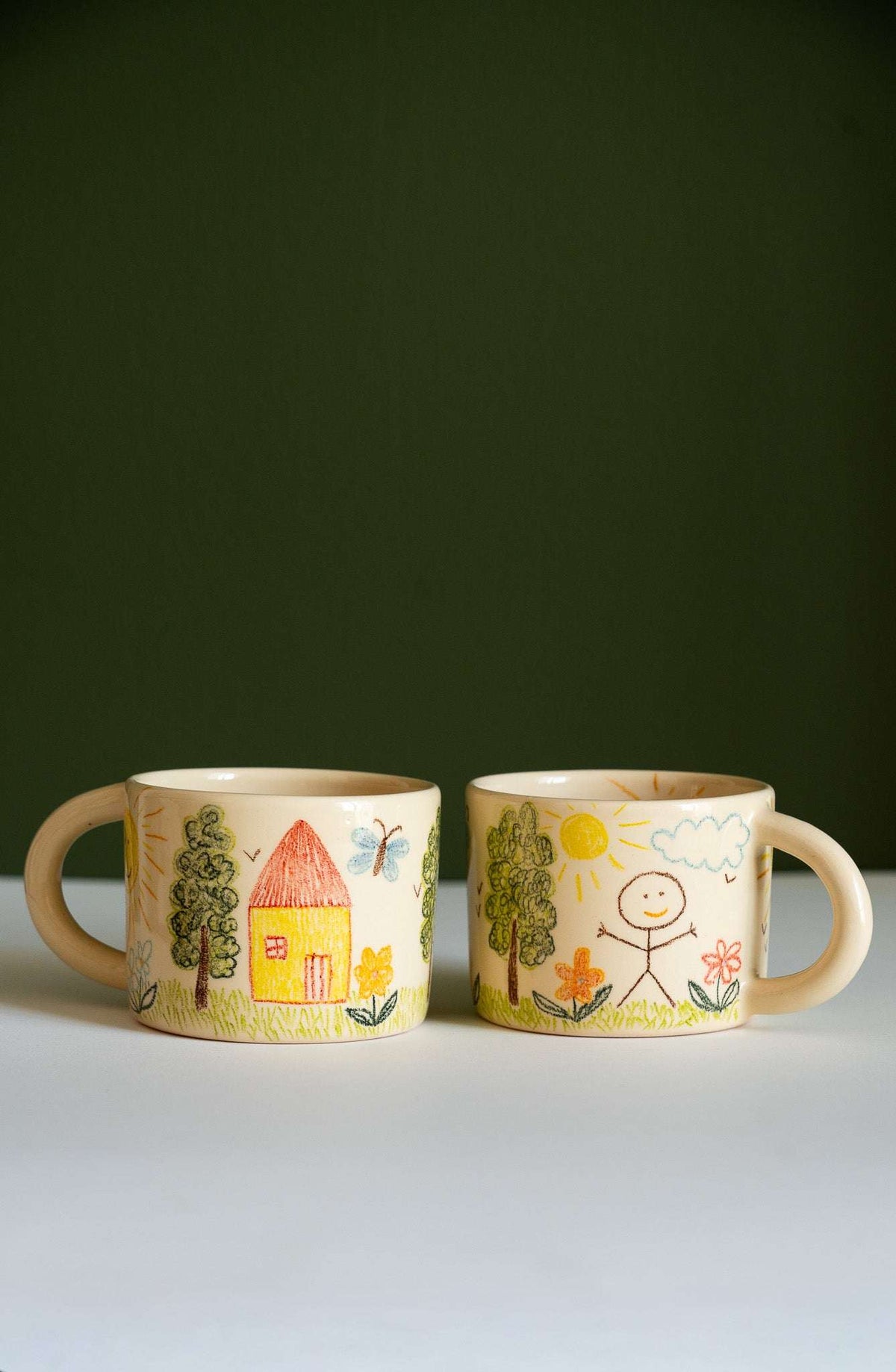 Kids Artwork Stoneware Mug in beige ceramic, hand-drawn crayon-style houses, trees, flowers, birds, and a smiling sun.