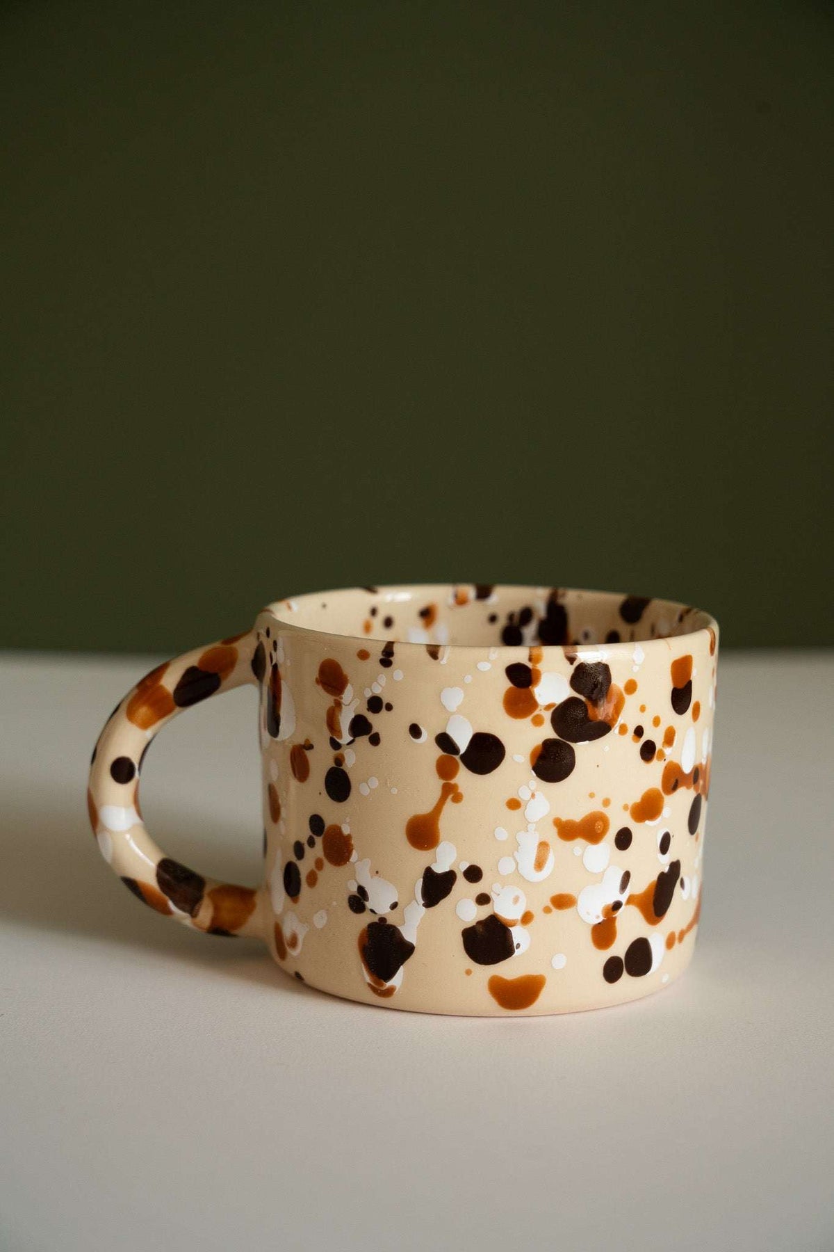 Splatter Stoneware Mug in glossy beige ceramic, covered with irregular brown, caramel, and dark espresso spots and a rounded handle.