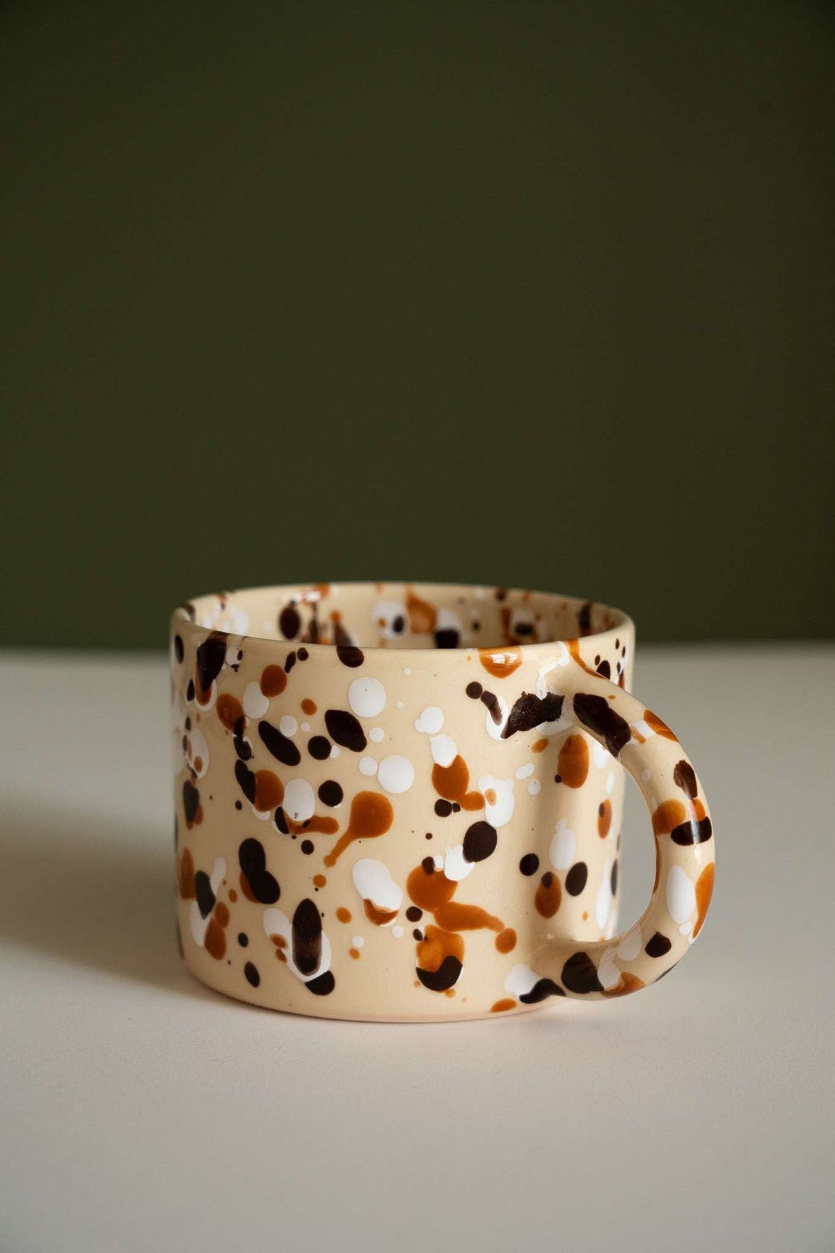 Splatter Stoneware Mug in glossy cream ceramic, hand-painted with irregular brown, black, and amber spots and a rounded handle.