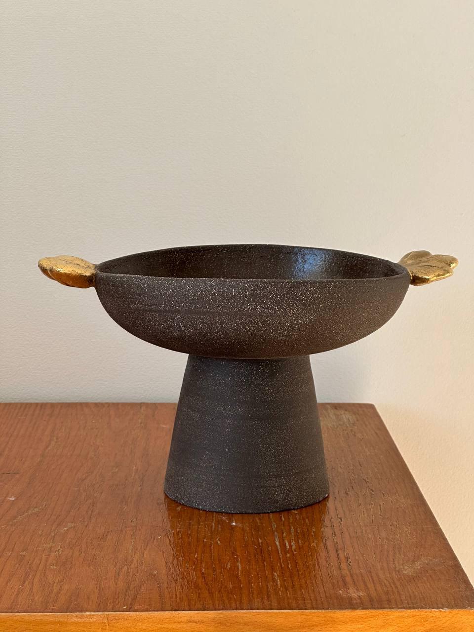 Pedestal fruit bowl in dark brown textured stoneware, with a wide basin, tall stem and gold leaf-shaped handles.