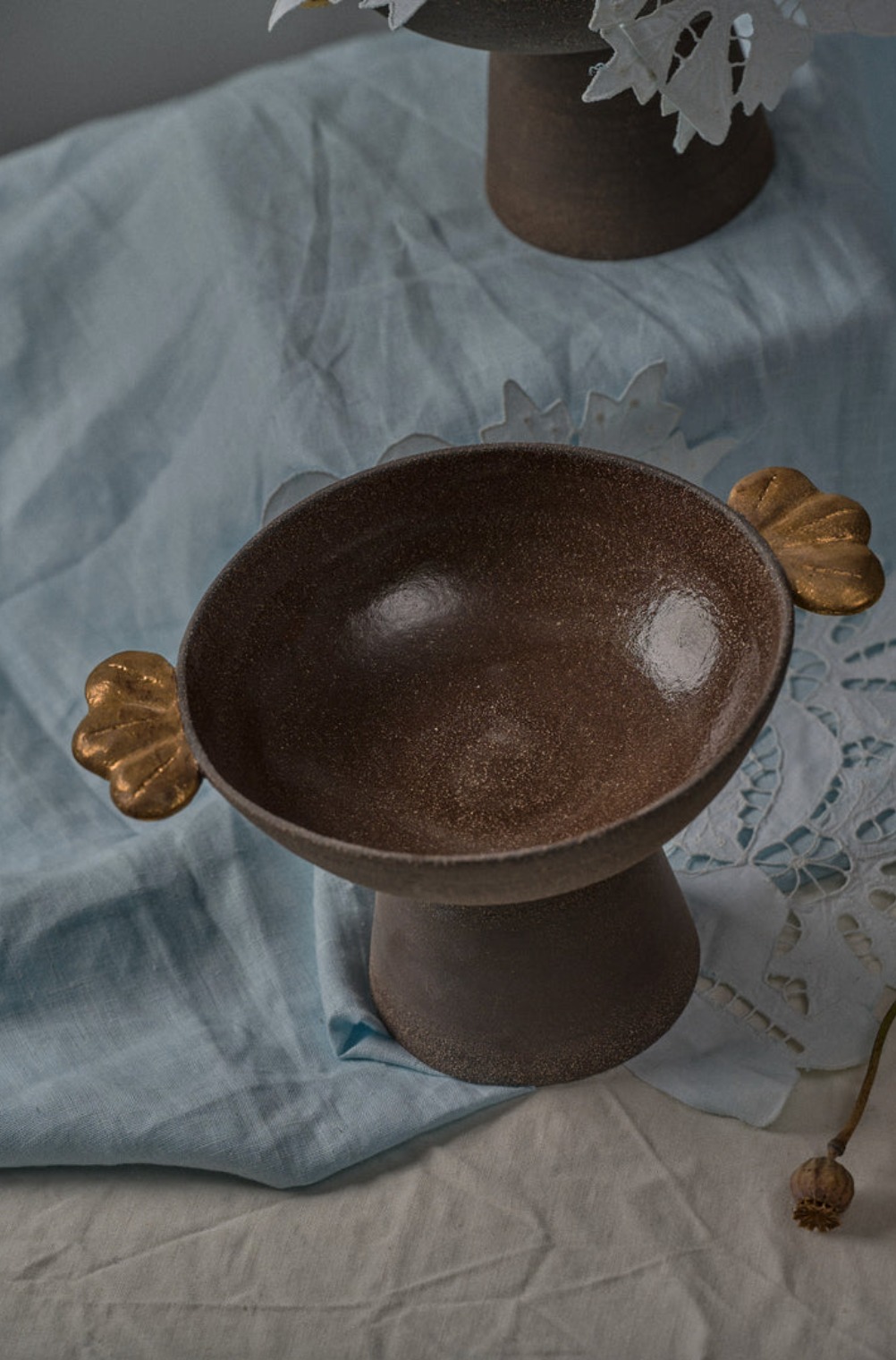 Pedestal fruit bowl in dark brown speckled stoneware, with a glossy interior and gold leaf-shaped handles.